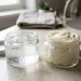 Two small glass jars side by side on a bathroom counter, one holding clear glycerin gel, the other a thick pale swirl of shea butter, with soft natural light showing their different t